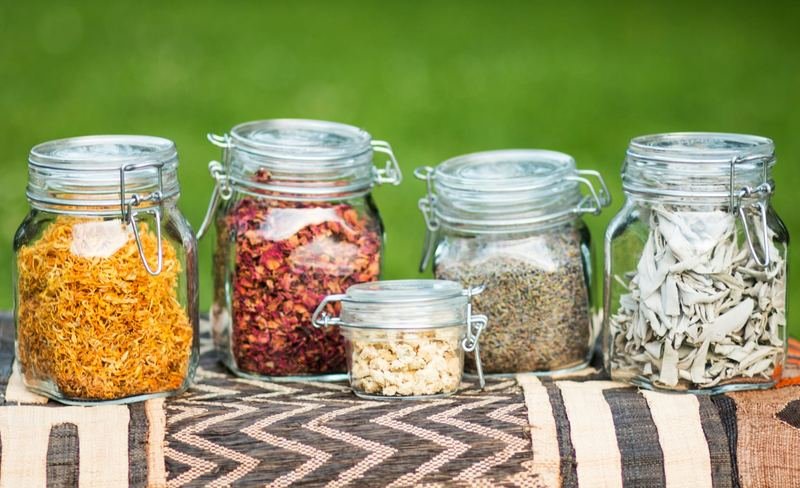 Bocaux de calendula, pétales de rose, lavande et sauge séchés, posés sur un tissu africain.