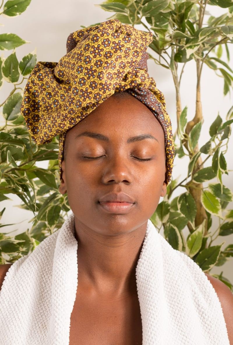 Une femme les yeux fermés, un foulard wax noué sur la tête, une serviette sur les épaules.