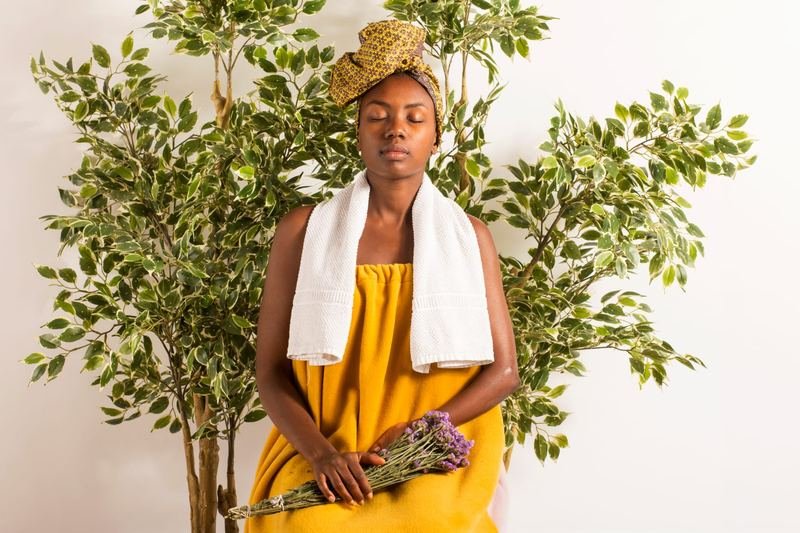 Une femme assise, enveloppée dans une cape jaune pour le bain de vapeur, tient un bouquet de lavande séchée.